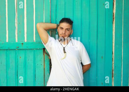 Young man in a white shirt and his hand behind his head standing next to a green wooden wall. Stock Photo