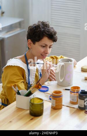 Young smiling creative woman working in her own cozy pottery studio, making ornament on ceramic jug Stock Photo