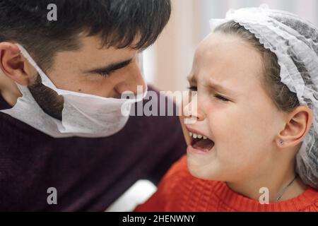 Portrait of dad in disposable mask on face and little tearful girl in medical disposable cap crying in pain after medical procedures in hospital Stock Photo