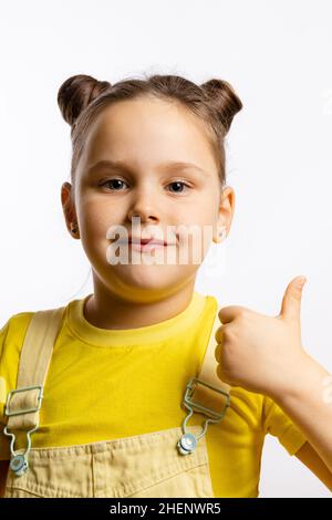 Portrait of charming little girl with shining bright smile with thumb up in yellow t-shirt and jumpsuit with two nice piggy tails on white background  Stock Photo