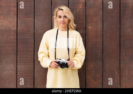Woman holding a camera while facing the camera and leaning on a wooden wall Stock Photo