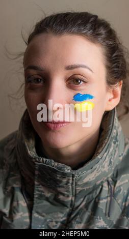 Indoor portrait of young girl with blue and yellow ukrainian flag on her cheek wearing military uniform, mandatory conscription in Ukraine, equality Stock Photo