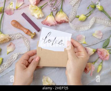 Hands with Card LOVE inside envelope near by pink flowers, wax seal and ribbons on a marble table top view. Romantic scene with handwritten card flat Stock Photo