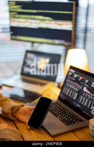 Cozy workplace with laptop and desktop computers with programming code on the screens. Programmer work table, Close-up view on phone with empty space Stock Photo