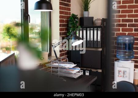 Close up of desk and bookshelf with decorations in office. Empty table with documents and computer, shelves with minimal decor. Water dispenser and bookcase with ornaments and plants. Stock Photo