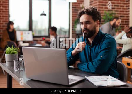 Doubtful pensive entrepreneur having uncertain think about marketing project analyzing company turnover on computer. Businessman working at business collaboration in startup office. Diverse team Stock Photo