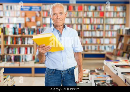 Intelligent older man holding out books to somebody while standing near racks Stock Photo