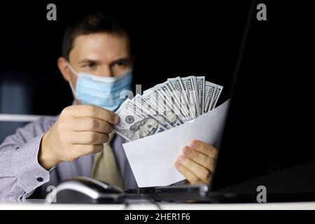 US dollars in male hands. Man in mask and office clothes pulls money out of an envelope sitting at laptop, wages, bonus or bribe concept Stock Photo