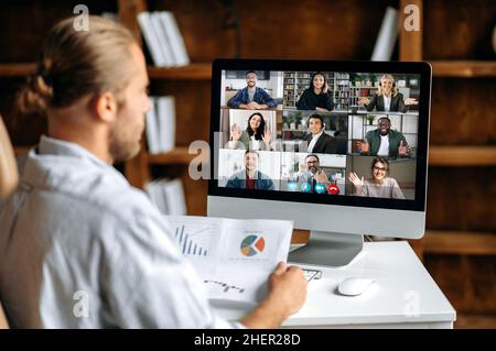 Online briefing, virtual business meeting. View over the shoulder of a company employee communicating with a group of multi-ethnic colleagues via video conference, analyzing financial statements Stock Photo