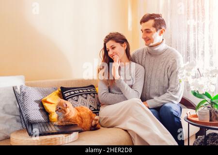 Young family couple calling through video chat with cat using laptop at home. Ginger pet sitting on couch looking at screen. Woman waving saying hi Stock Photo