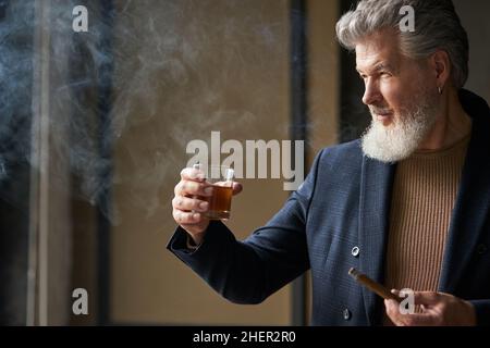 Portrait of sophisticated mature businessman with beard looking away while holding glass of whisky and cigar, standing in loft interior Stock Photo