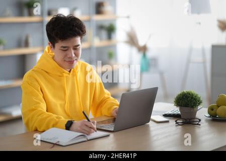Asian man sitting at desk, using pc writing in notebook Stock Photo