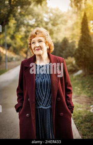 A cute mature woman in autumn stands outdoors in an unbuttoned red coat and a blue dress with short hair. Smiles. Stock Photo
