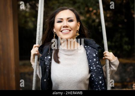 Close-up portrait of a young cheerful tatar woman swinging on a swing in the forest in autumn. High quality photo Stock Photo