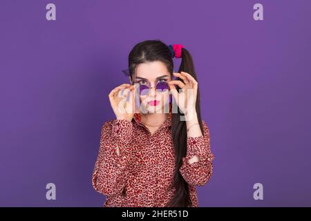 Young pretty woman dressed in 90s style looks with surprise at the camera holding round glasses. Stock Photo
