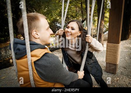 Cheerful young mixed race couple looking at each other while sitting on a swing outdoor. Stock Photo