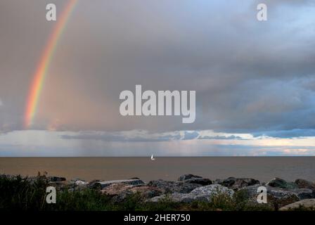 Distant, solitary sailing boat on a calm sea with a rainbow in the dark clouds above and boulders on the shore, Norfolk coast, UK Stock Photo
