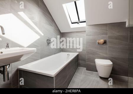 Cowlinge, Suffolk, England - 28 March 2019: Luxury generic sunlit bathroom suite with grey tiles and white painted sloping ceiling. Stock Photo