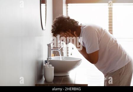 Young indian man washing face with clear water from tap bending over sink, doing skincare facial routine in bathroom Stock Photo