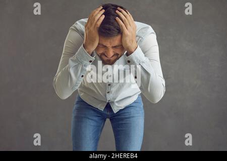 Upset frustrated man holding his head in despair standing against grey studio background Stock Photo