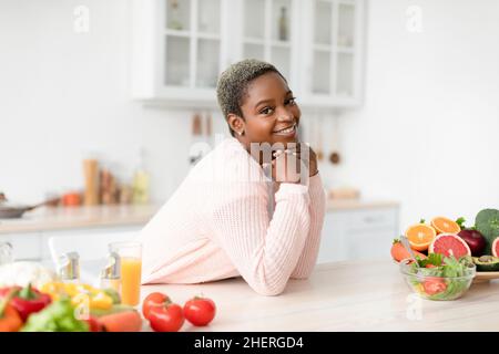 Happy young african american female blogger look at camera in scandinavian kitchen interior with table with vegetables Stock Photo