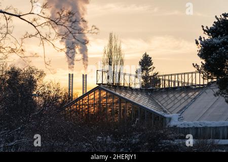 Greenhouse building in winter garden against smoking chimneys of boiler plant at sunset Stock Photo