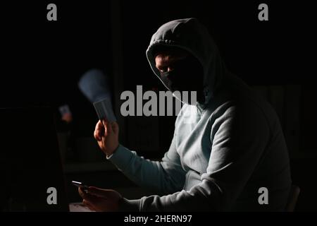 Hacker in dark jacket looking at laptop screen in dark room is holding bank card Stock Photo