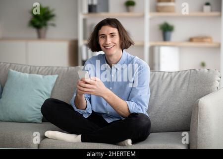 Enjoying weekend. Happy young guy using smartphone while sitting cross legged on couch at home, copy space Stock Photo