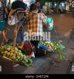 Street vendor starting her work. Backview of a woman carrying a shoulder pole with fresh fruits. Straw conical hat. City lights on. Hanoi, Vietnam. As Stock Photo