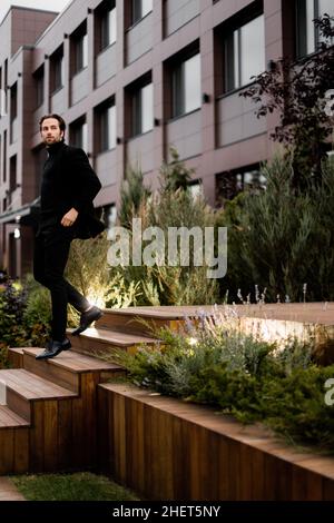 Stylish dressed businessman is walking down by stairs, looking at the side, posing for photo outdoor. Successful handsome male in classic suit showing Stock Photo