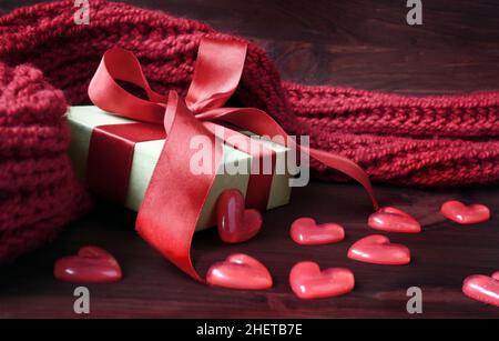 White gift box and plastic hearts with red knitted scarf on red wooden background. Holiday concept. Stock Photo