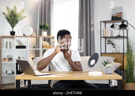 Young athletic African man shaves the beard on the cheek with an electric razor looking in the mirror. The concept of spending free time at home. Hygiene, health and beauty Stock Photo