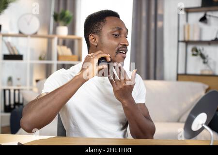 Young athletic African man shaves the beard on the cheek with an electric razor looking in the mirror. The concept of spending free time at home. Hygiene, health and beauty Stock Photo
