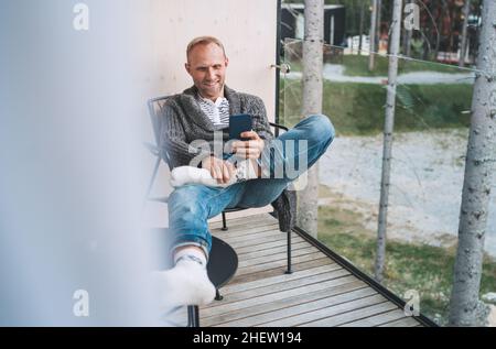 Smiling middle-aged man dressed open cardigan, jeans, and warm socks sitting on forest house balcony and using modern smartphone. Everyday lifestyle p Stock Photo