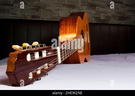 classical guitar lying on the bed in front of a brown leather back and a stone wall Stock Photo