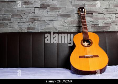 classical guitar lying on the bed in front of a brown leather back and a stone wall Stock Photo