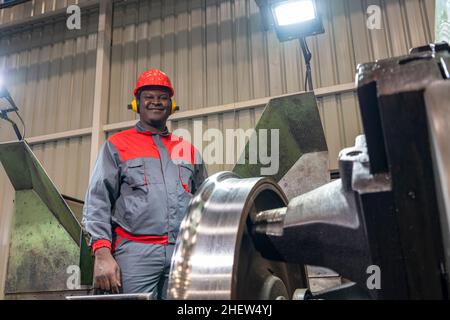 Portrait Of Black Machine Operator In Red Helmet, Hearing Protectors And Work Uniform Next To Lathe Machine. Stock Photo