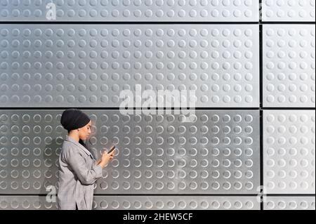 Side view of a muslim woman standing using her phone with a wall of iron panels with circles patterns. Stock Photo