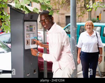 Positive African American man buying ticket for street parking in modern parking meter Stock Photo