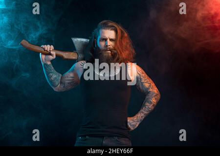 Handsome man with cigar holds old axe. Bearded lumberjack. Serious male with tattoos with long hair. Studio shot Stock Photo