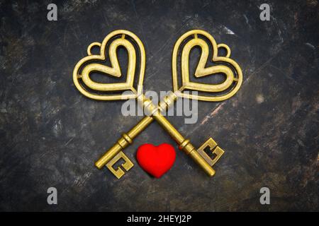 Crossed old-fashioned brass keys with a small red heart on a dark grungy background. Romantic symbol of expressing a special trust and commitment. Stock Photo