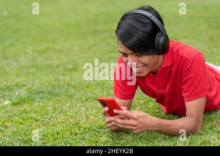 Cheerful smiling young latino man lying on grass with smartphone. Stock Photo