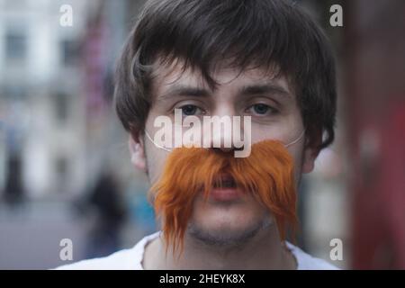 Portrait of a young man with fake red mustache. Mustache does not grow, but he wants to have, concept. Stock Photo