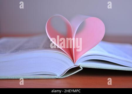 An unfolded book on a table with a pink heart made of pages. Blurred background. Copy space. Stock Photo