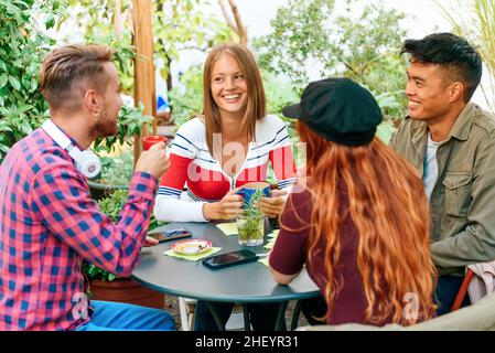 Group of happy young diverse male and female friends in casual clothes smiling and looking at each other while drinking coffee together on terrace of Stock Photo