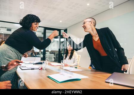 Cheery businesswomen fist bumping each other before a meeting in a boardroom. Two colleagues smiling cheerfully while greeting each other. Group of bu Stock Photo