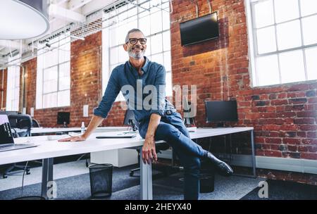 Mature businessman smiling while sitting in a modern workplace. Happy mature businessman sitting alone on an office desk. Experienced businessman feel Stock Photo