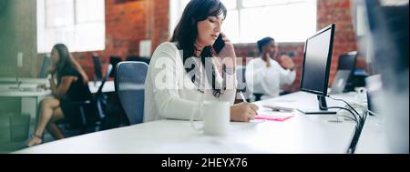 businesswoman making notes while speaking on a phone call. Creative businesswoman connecting with her business clients in a modern office. Young busin Stock Photo