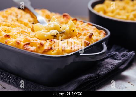 Classic mac and cheese with basil on top and mac and cheese baked in oven in background with forks and raw macaroni on a rusticžwooden background Stock Photo