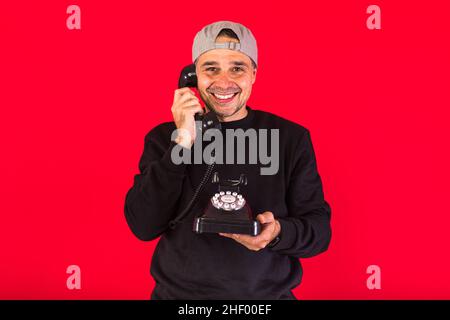 Man in a cap dressed in black, answers a call with a retro telephone from the 60s and smiles, on a red background, with copy space. Concept of contact Stock Photo